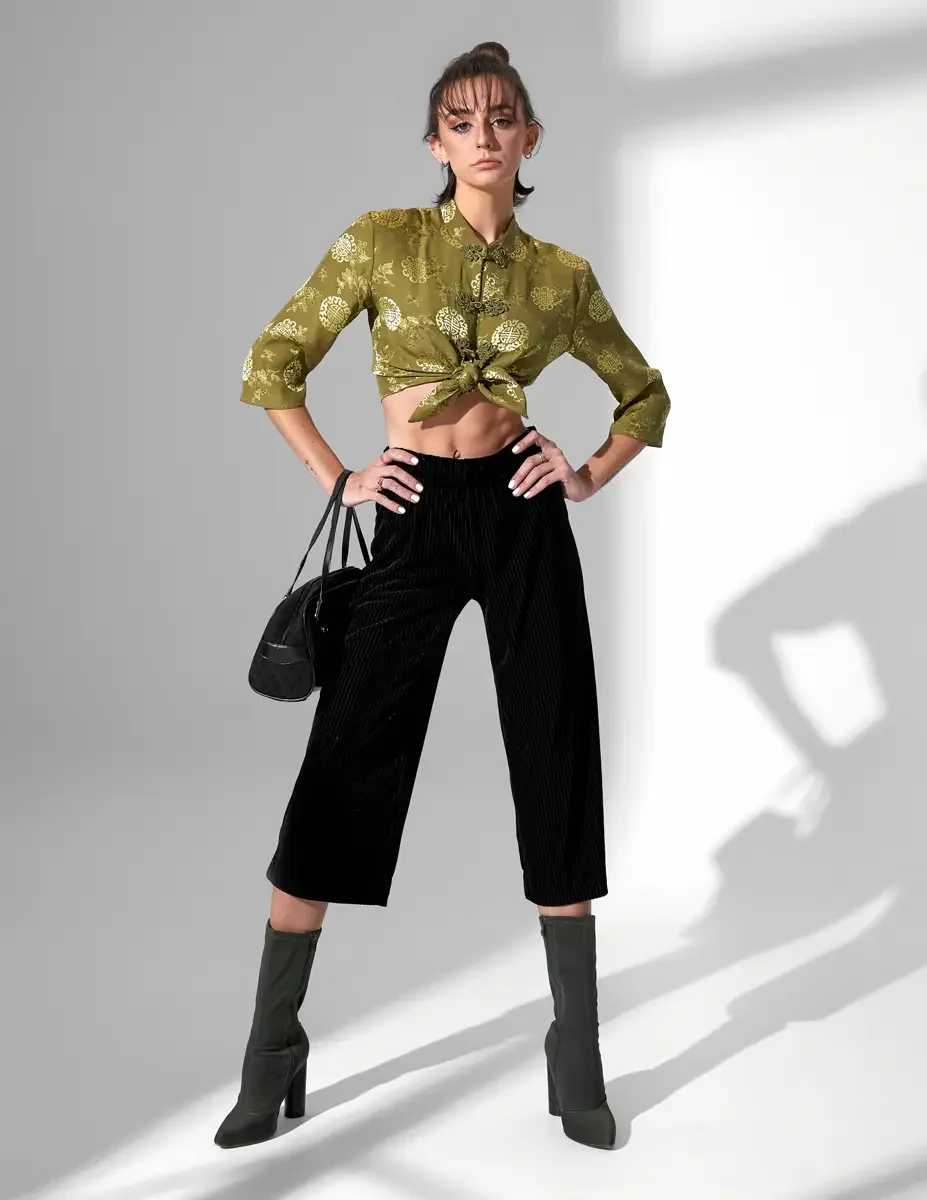 Model Macy, full length, hands on hips, olive green satin jacquard tied crop top with gold medallion print and frog closures, black wide-leg ribbed velvet culottes, olive green block-heel sock boots, black suede barrel bag, New York studio test shoot