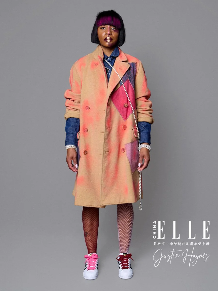 A woman with a black bob haircut with purple bangs, wearing a colorful oversized coat over a denim shirt, fishnet stockings, and sneakers, standing against a gray background for a fashion magazine cover.