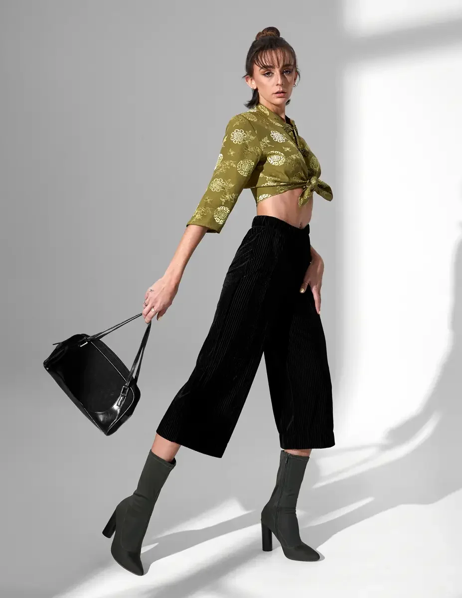 
Alt text: Macy, full length, mid-stride, olive green satin jacquard tied crop top with gold medallion print and frog closures, black wide-leg ribbed velvet culottes, olive green block-heel sock boots, black suede barrel bag, New York studio test sho