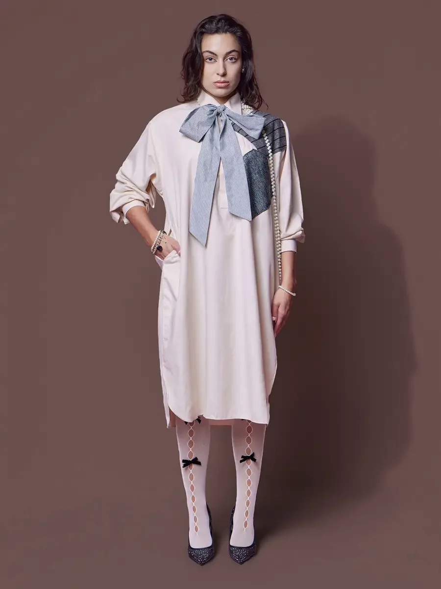 JUS10H 2025 content library, model in blush oversized shirt-dress with striped bow and metallic herringbone patchwork panel, long pearl necklace, bow-detail sheer tights and glitter pumps, hand on hip, brown background, photographed by Scott Parker P