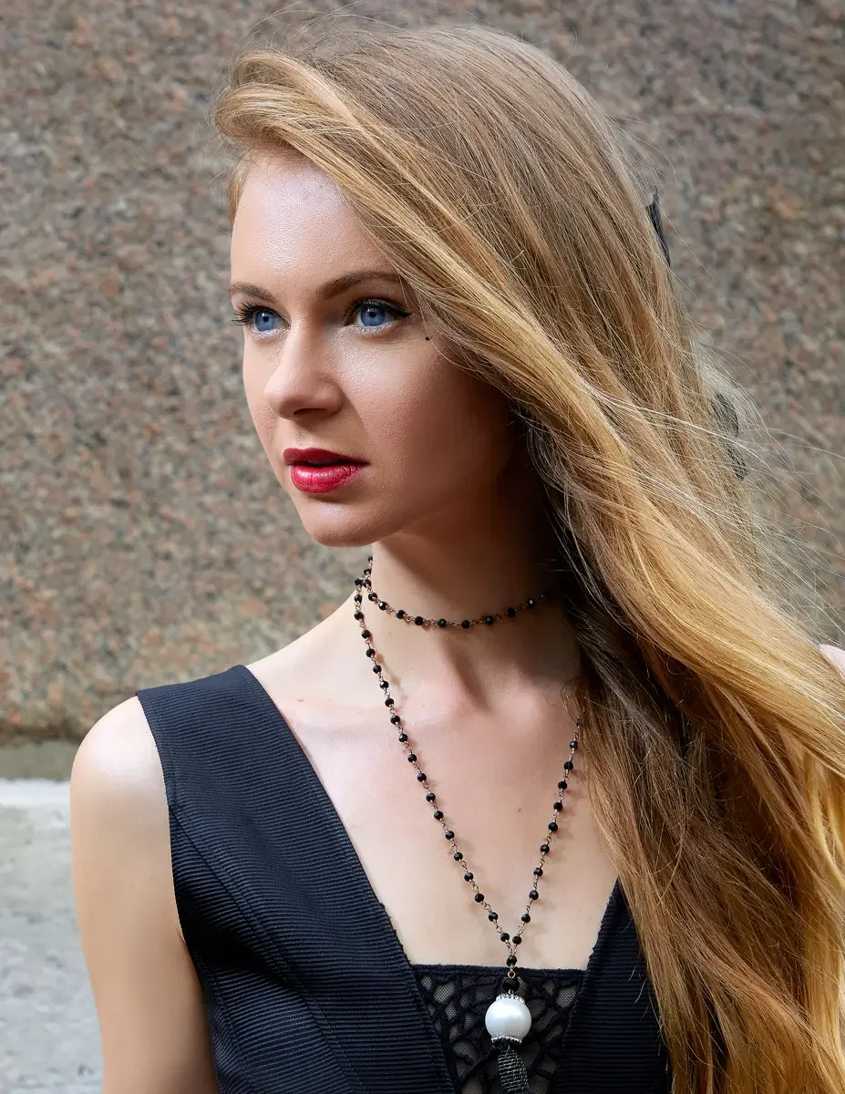 Julia, close-up portrait, black bandage-style sleeveless gown with deep V-neckline and mesh lattice bodice detail, layered black bead choker and long rose gold chain pendant necklace with baroque pearl drop and tassel, Upper East Side park test shoot