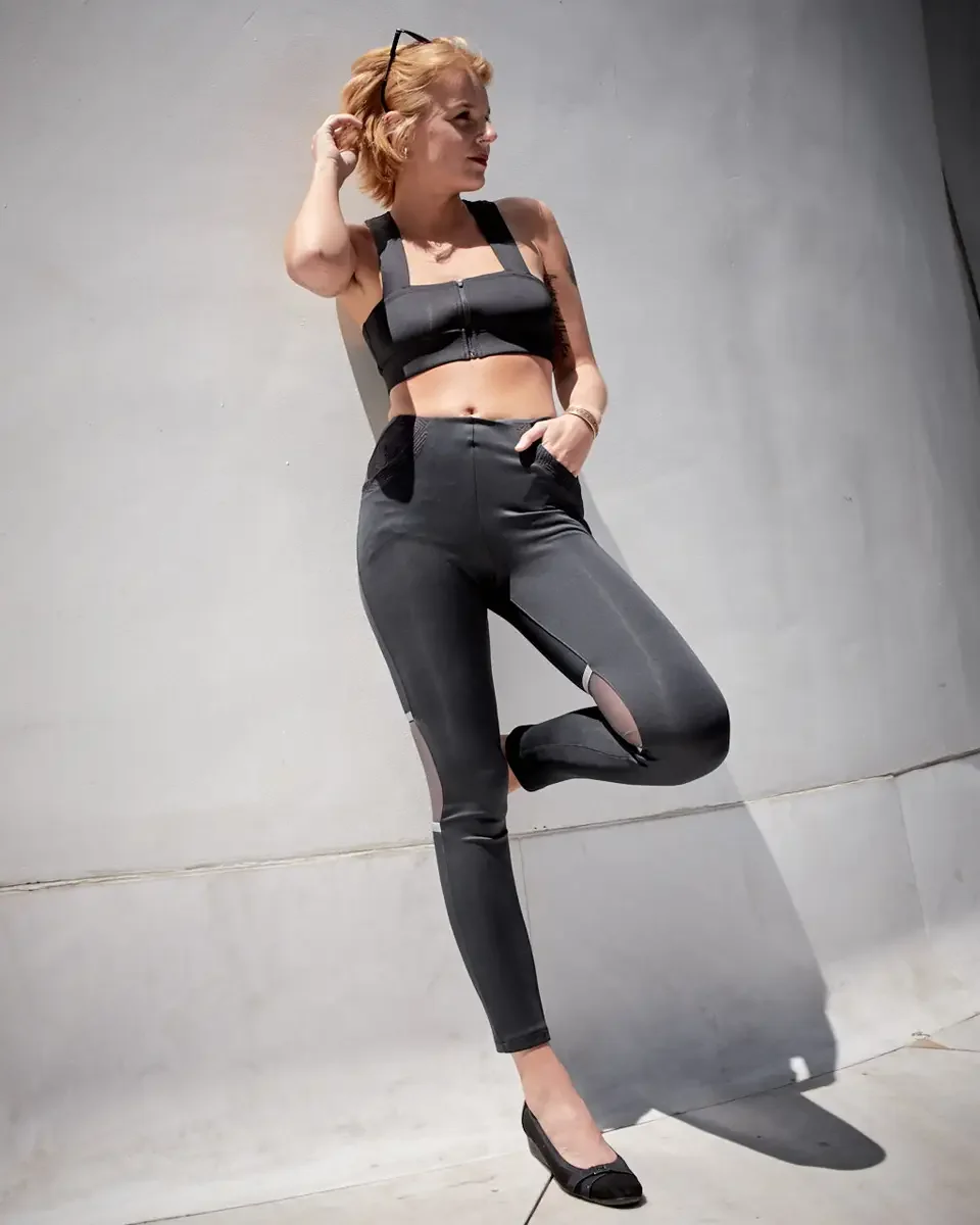 Satine, full length, leaning against concrete wall, one knee raised, black zip-front structured crop top, black high-waist leggings with sheer mesh panel inserts and reflective stripe, black flat loafers, gold bangle, Battery Park area test shoot, ph