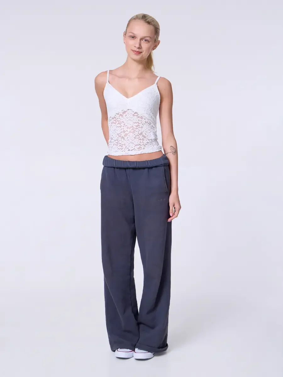 Model portfolio development session, commercial look, white lace crop camisole top and wide-leg charcoal sweatpants with elastic waistband, full-length, facing camera, arms relaxed at sides, soft smile, white Converse sneakers, light gray background,