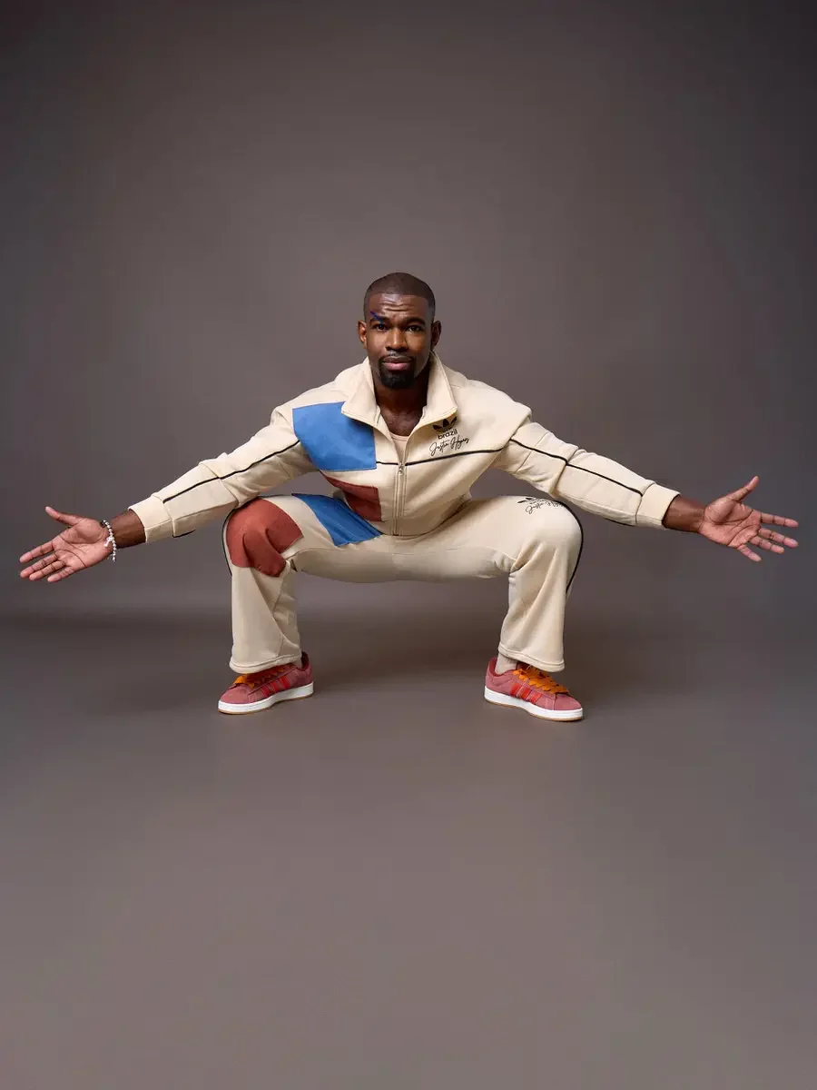 JUS10H x Adidas Brasil campaign, model in wide squat with arms extended, cream color-blocked tracksuit with blue and terracotta suede panels, Adidas Campus 00s in pink suede with red stripes and orange laces, warm brown background, photographed by Sc