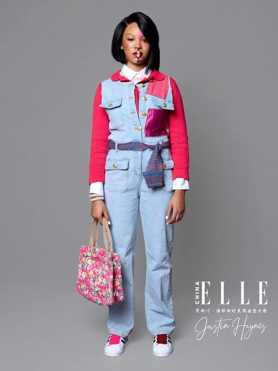Fashion model standing against a plain gray background wearing a year tech denim jumpsuit with pink sleeves, pink patch on chest, and plaid belt. She holds a pink floral tote bag and wears white sneakers with pink accents. She has black hair styled i