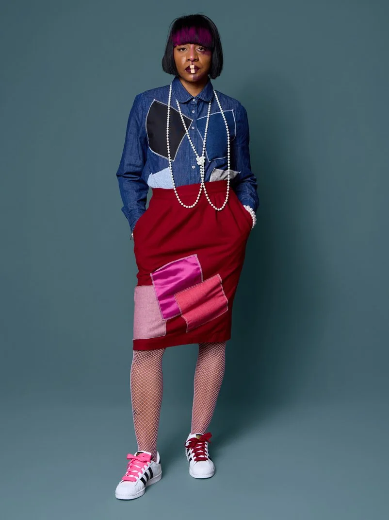 A woman standing with her hands in her pockets, wearing a patchwork denim shirt, a red skirt with pink patches, fishnet stockings, white sneakers with pink and black accents, and layered pearl necklaces. She has black hair with pink highlights and da