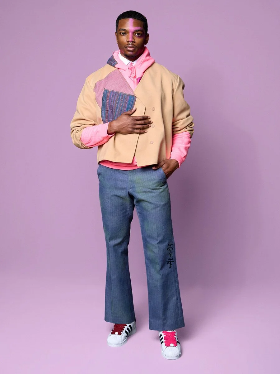 A young man wearing a pink hoodie, a beige jacket, blue jeans, and white sneakers with pink and black accents stands against a purple background. He has his right hand on his chest and his left hand in his pocket.