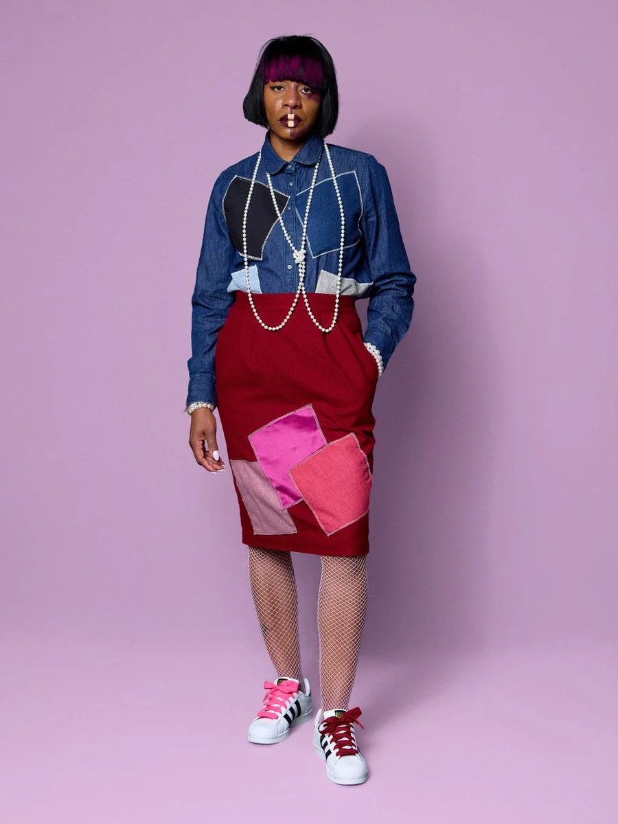 Woman with black hair and purple bangs wearing a denim shirt with pearl necklaces, a red patchwork skirt, fishnet stockings, and white sneakers with pink and red laces, standing against a purple background.