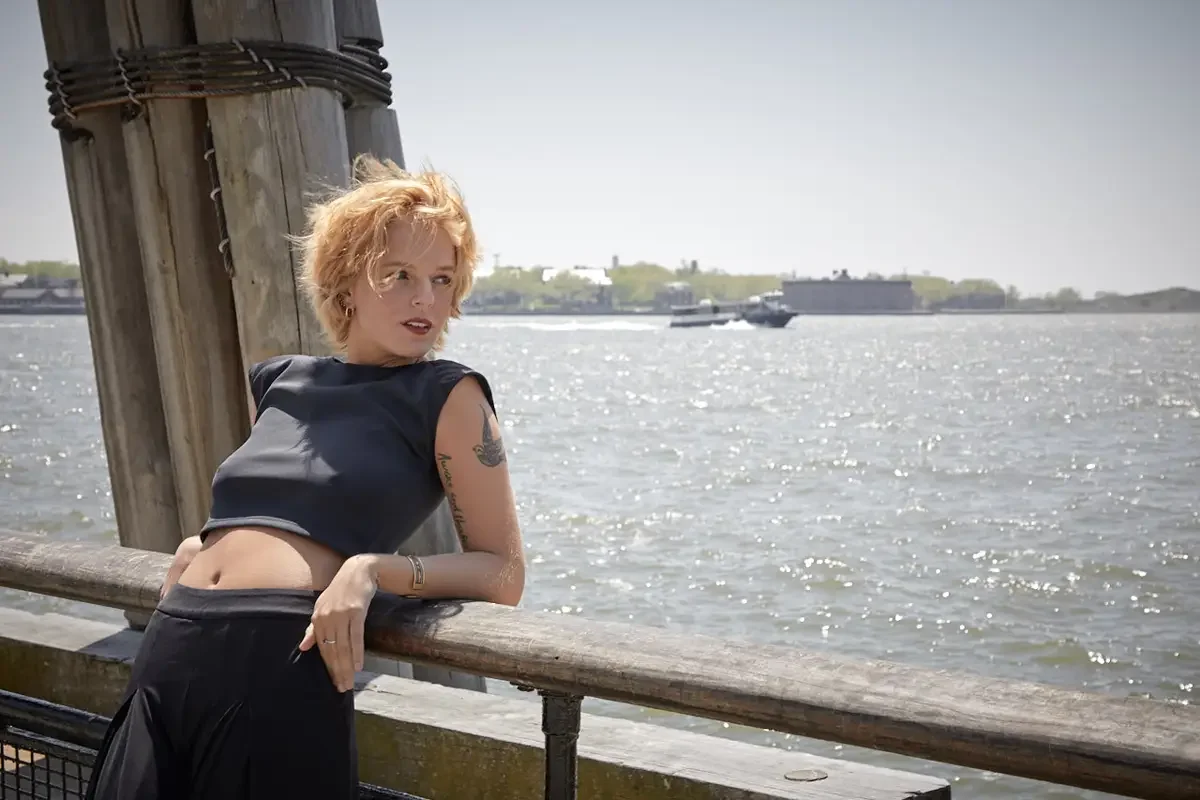 Satine, three-quarter crop, leaning on wooden pier railing, black structured cap-sleeve crop top, black wide-leg palazzo pants, gold bangle, bird and script tattoo on upper arm, Ellis Island visible in background, Battery Park waterfront test shoot, 