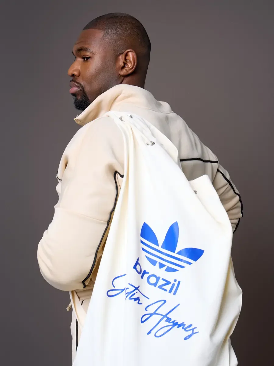  JUS10H x Adidas Brasil campaign, three-quarter rear portrait, model in cream tracksuit with black piping carrying white JUS10H Adidas Brasil drawstring bag with blue trefoil logo and Justin Haynes signature, warm brown background, photographed by Sc