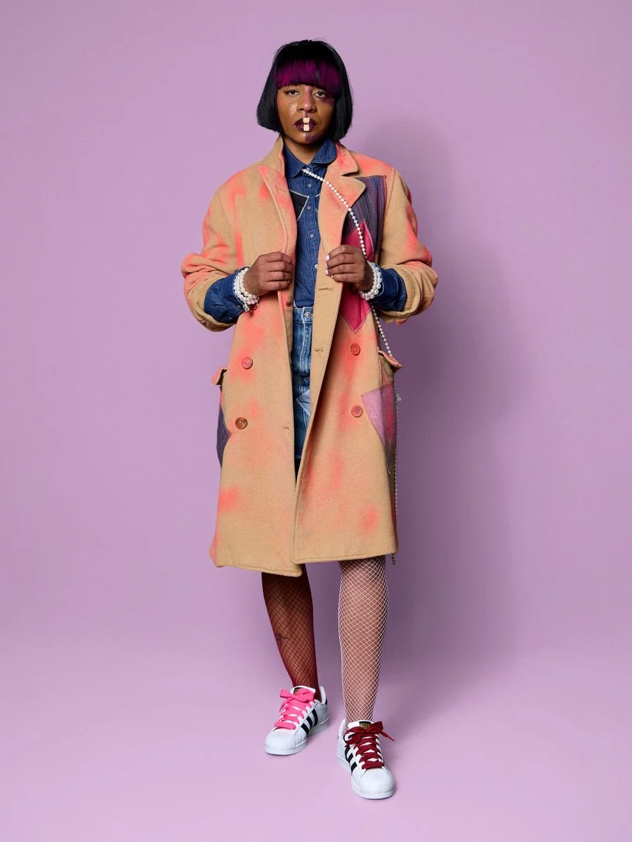 A woman with purple and black hair wearing a long, pinkish-beige coat with patches, a denim shirt, fishnet stockings, and white Adidas sneakers with black stripes, standing against a purple background.