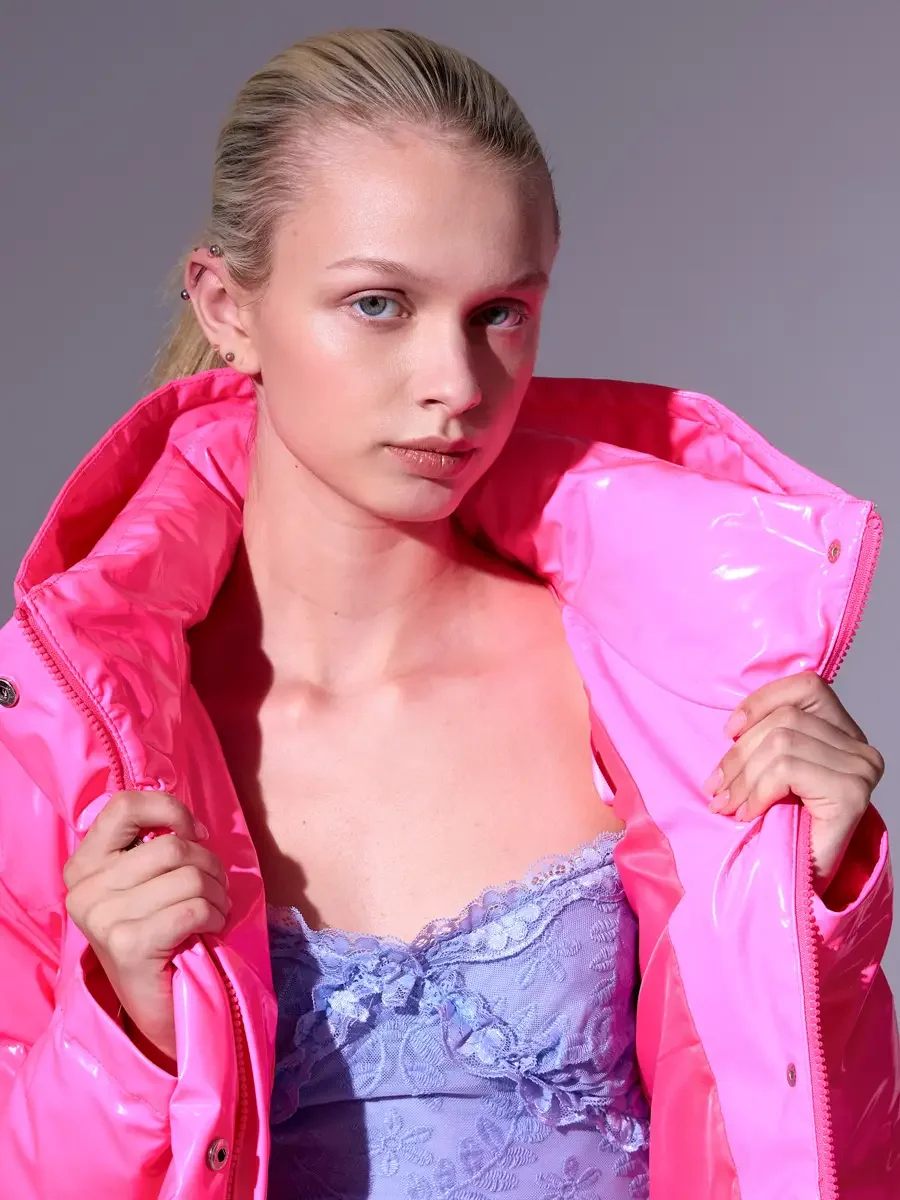 Model portfolio development session, high fashion editorial look, hot pink vinyl puffer jacket held open over periwinkle blue lace asymmetrical midi dress, close-crop, both hands pulling lapels wide, direct gaze, pink ambient light cast across face f