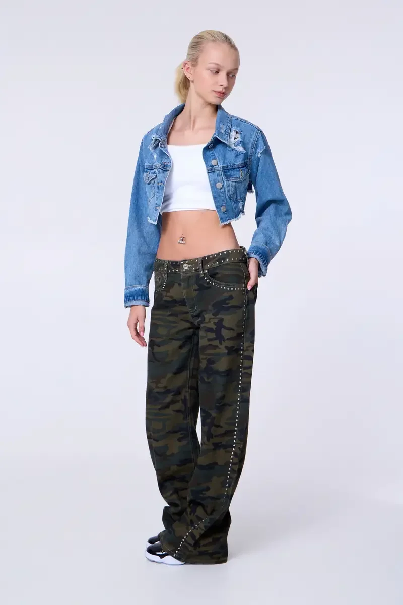 Model portfolio development session, commercial look, cropped distressed blue denim jacket over white crop tank, low-rise studded camouflage wide-leg trousers with silver stud trim at waistband and side seam, full-length, slight turn to camera, one h