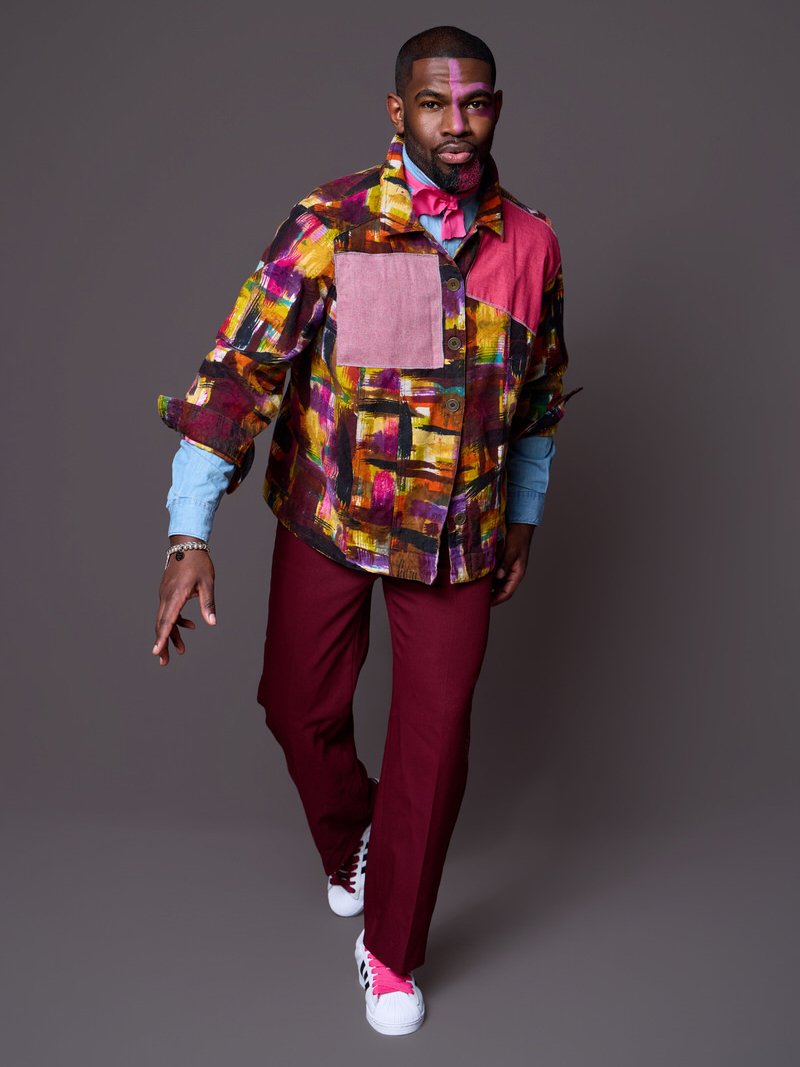 A man wearing a colorful abstract patterned jacket, maroon pants, white sneakers with black and pink accents, and a light blue shirt with rolled-up sleeves, standing against a plain gray background.