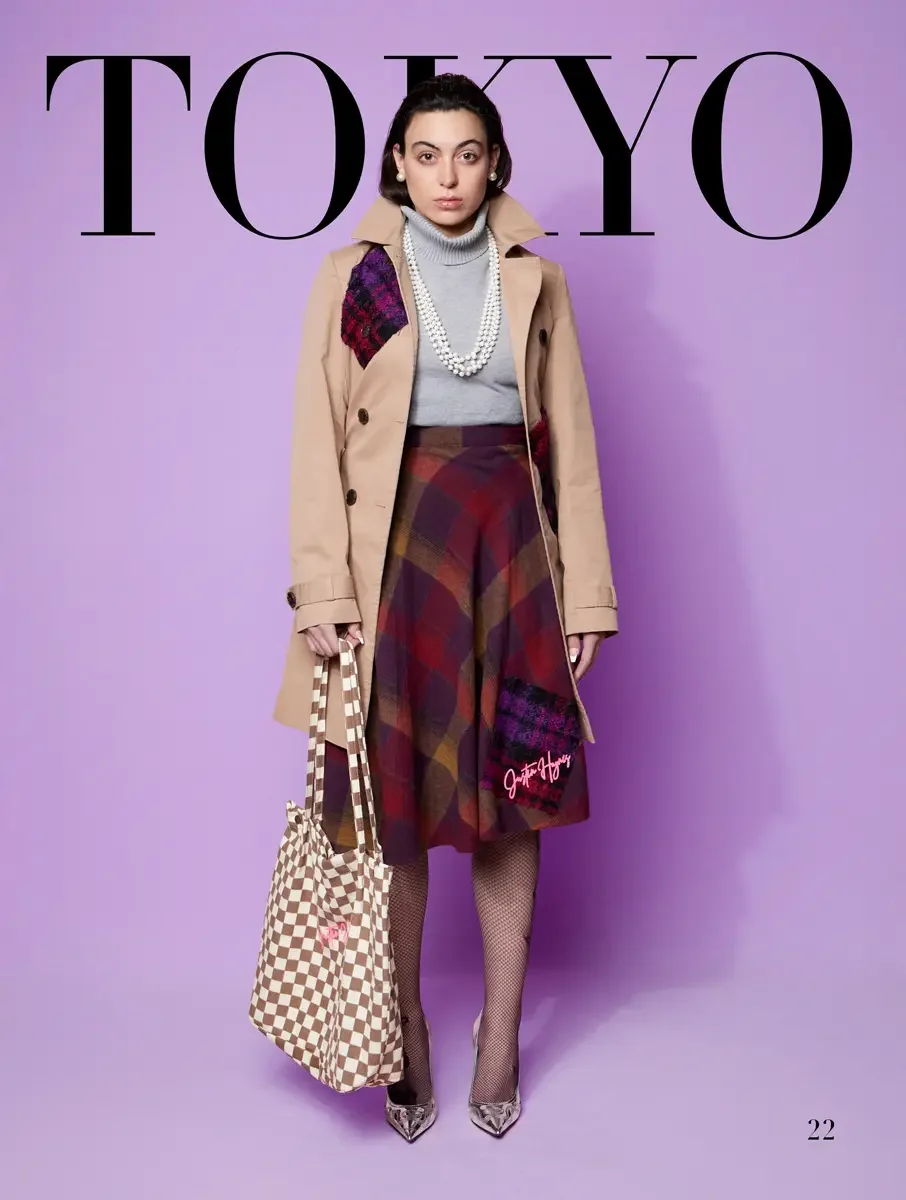 JUS10H 2025 Tokyo lookbook, short tan trench coat with cranberry purple tartan patch over grey turtleneck and large plaid midi skirt with JUS10H signature detail and checkered tote bag, lavender background, photographed by Scott Parker Photo for the 