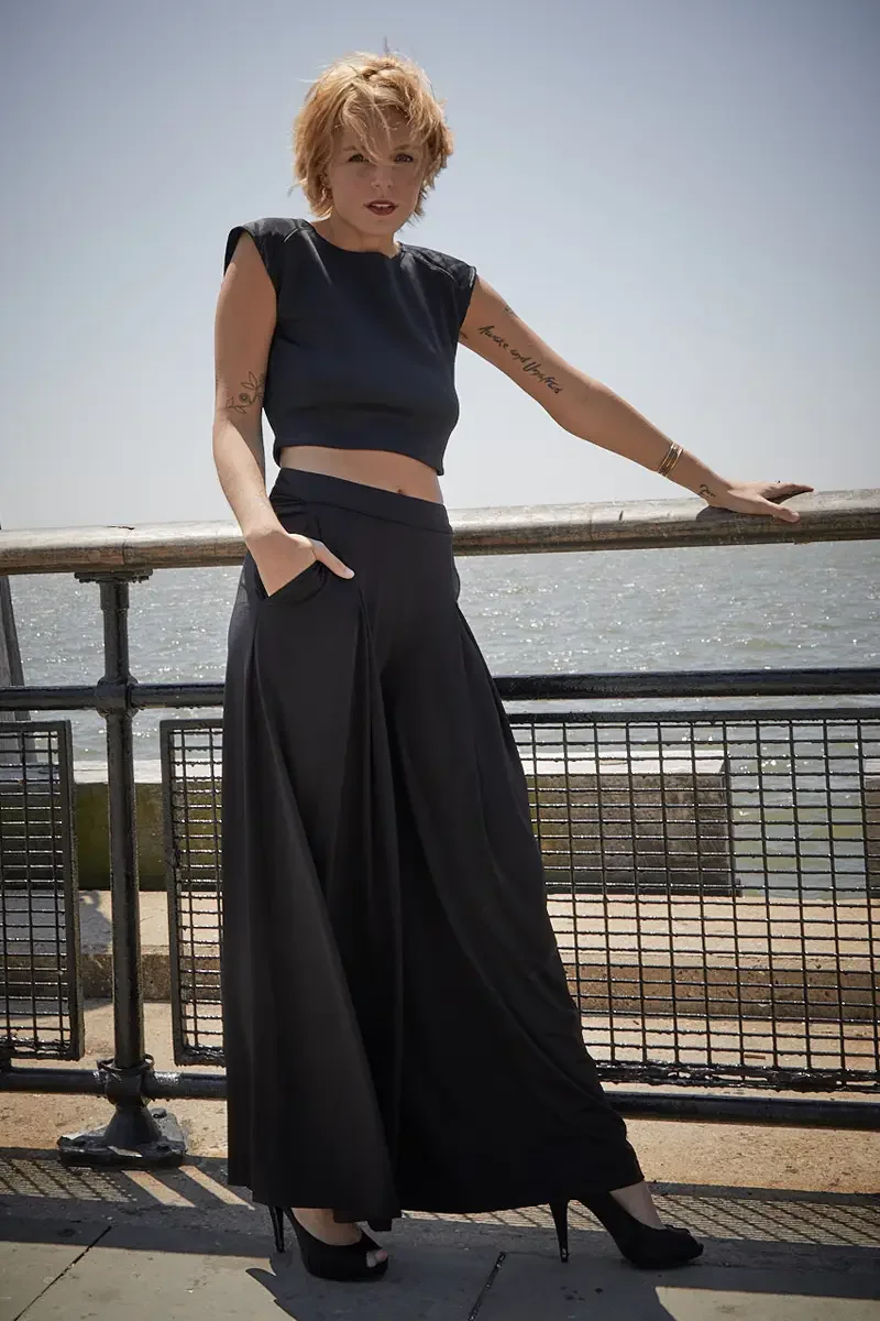 Satine, full length, hand in pocket, leaning on waterfront railing, black structured cap-sleeve crop top with shoulder detail, black wide-leg pleated palazzo pants with wide waistband, black platform peep-toe heels, script and bird tattoo on right ar