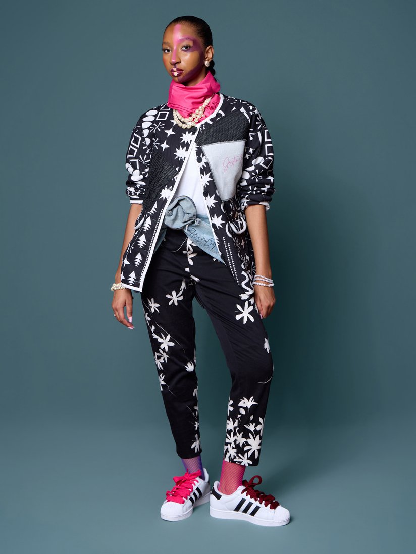 A woman standing against a plain background, wearing a black and white patterned outfit with a pink neck scarf, colorful makeup, jewelry, and sneakers.