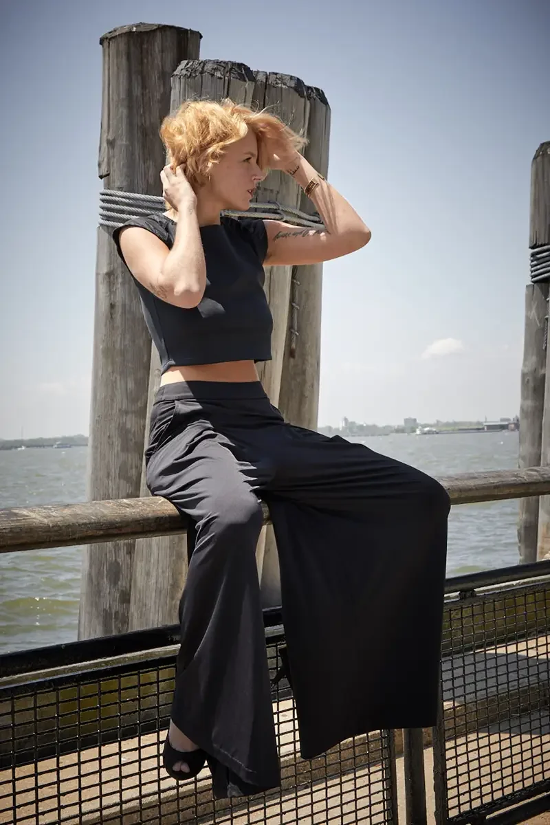 Satine, full length, seated on pier railing, hands at hair, black structured cap-sleeve crop top, black wide-leg pleated palazzo pants, black platform peep-toe heels, script tattoo on right arm, gold bangle, Battery Park pier test shoot, photographed