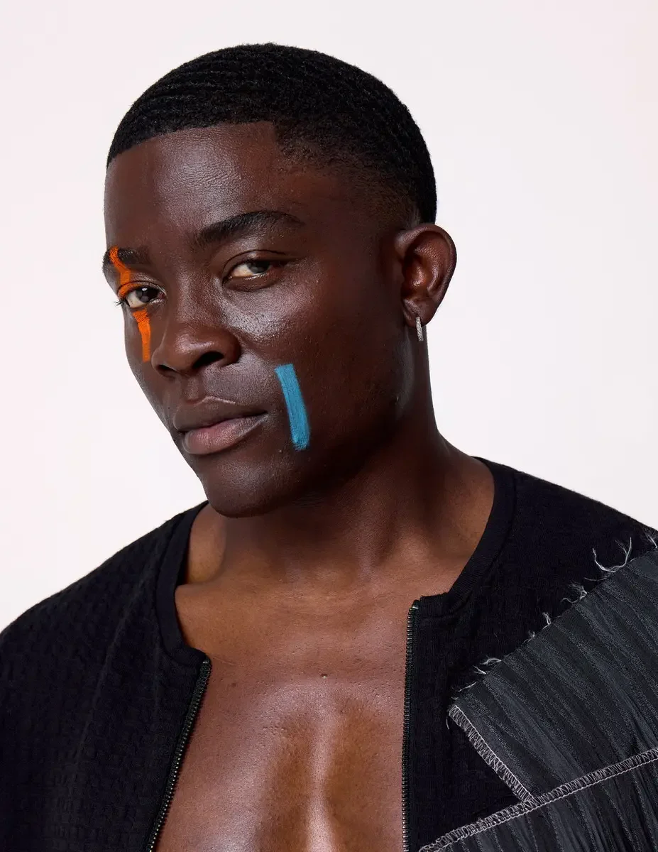 JUS10H Black Summer Jacket editorial, close portrait of male model in black collarless jacket worn open with gray patchwork panel, no shirt, white background, photographed by Scott Parker Photo