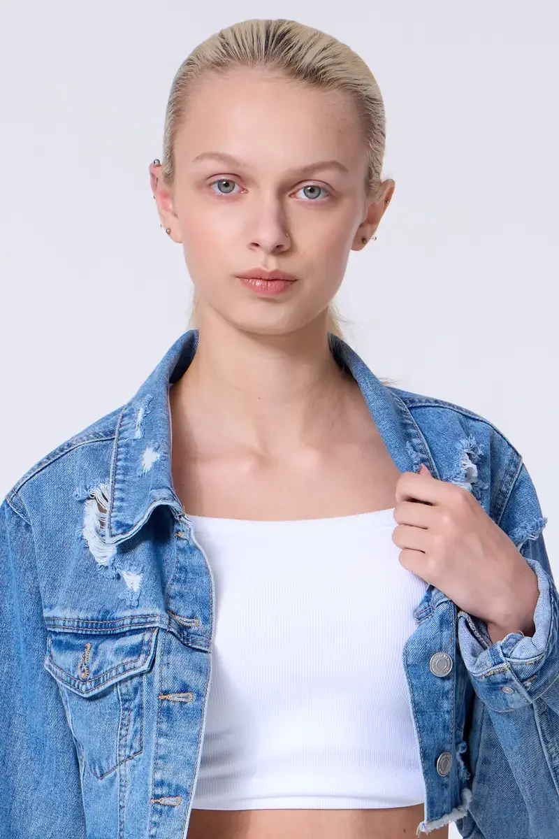 Model portfolio development session, commercial look, cropped distressed blue denim jacket over white crop tank, close-crop, hair pulled back sleek, one hand at jacket collar, direct gaze, distressed denim texture and collar detail visible, light gra