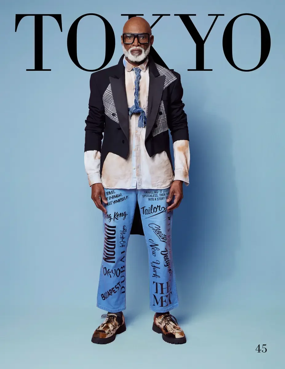 JUS10H 2025 Tokyo lookbook, a male model wearing a deep navy blue double-breasted blazer with houndstooth and plaid patchwork lapel panels over a white and tan distressed button-down shirt, light blue jeans hand-lettered with city names and travel qu