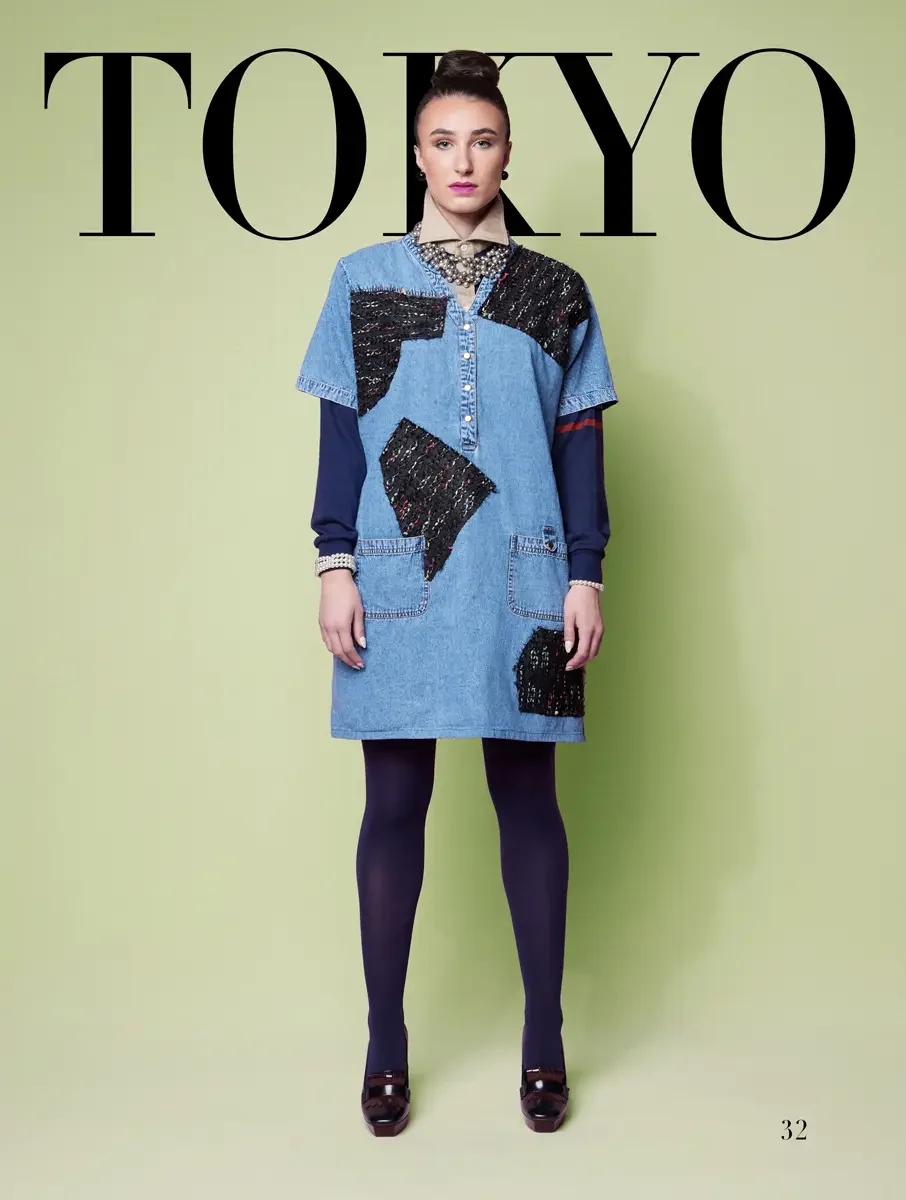 JUS10H 2025 Tokyo lookbook, a model wearing an oversized light-wash denim mini dress with bold black textured patchwork appliqués and patch pockets, layered over a navy long-sleeve top with pearl cuff accents, styled with a pearl collar necklace, nav