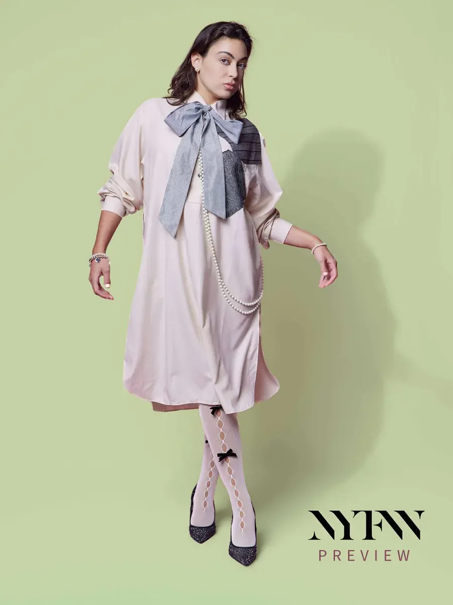 JUS10H 2025 print advertising, model in motion in blush oversized shirt-dress with striped bow, metallic and herringbone patchwork panel, long pearl necklace, bow-detail sheer tights and glitter pumps, sage green background, photographed by Scott Par