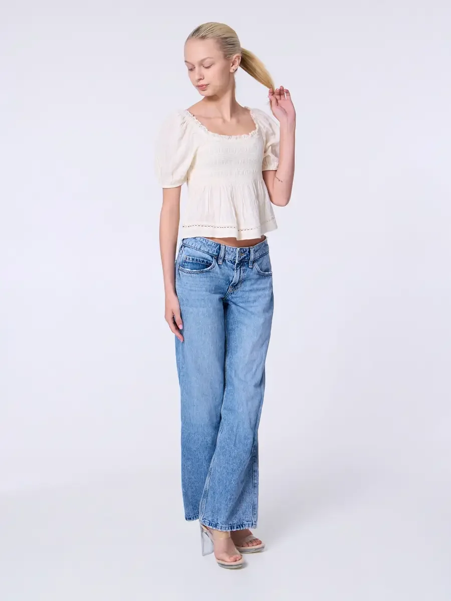 Model portfolio development session, commercial look, cream smocked square-neck puff-sleeve crop top and low-rise light wash wide-leg jeans, full-length, facing camera, one hand raised pulling ponytail, gaze downward, relaxed movement, nude heeled sa