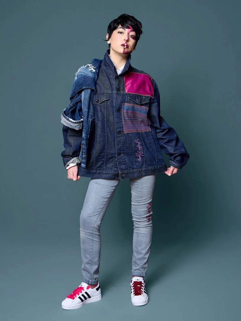 A person with short dark hair and pink highlights, wearing a denim jacket with colorful patches, light gray jeans with red embroidery, and white sneakers with red and black accents poses against a solid background.