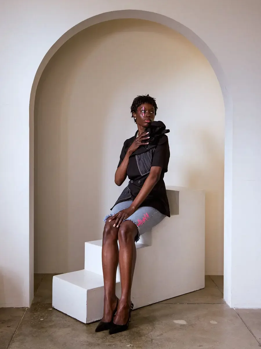 JUS10H Black Summer Jacket content library, female model in black blazer with oversized floral corsage and gray denim patchwork panel, seated on white steps inside arch, denim shorts with JUS10H logo, black pointed-toe heels, warm neutral background,