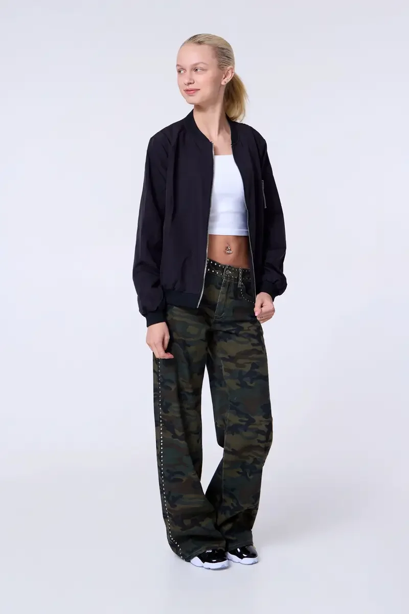 Model portfolio development session, commercial look, black bomber jacket open over white crop tank, low-rise studded camouflage wide-leg trousers with silver stud trim at waistband and side seam, full-length, three-quarter turn, gaze off-camera, sof