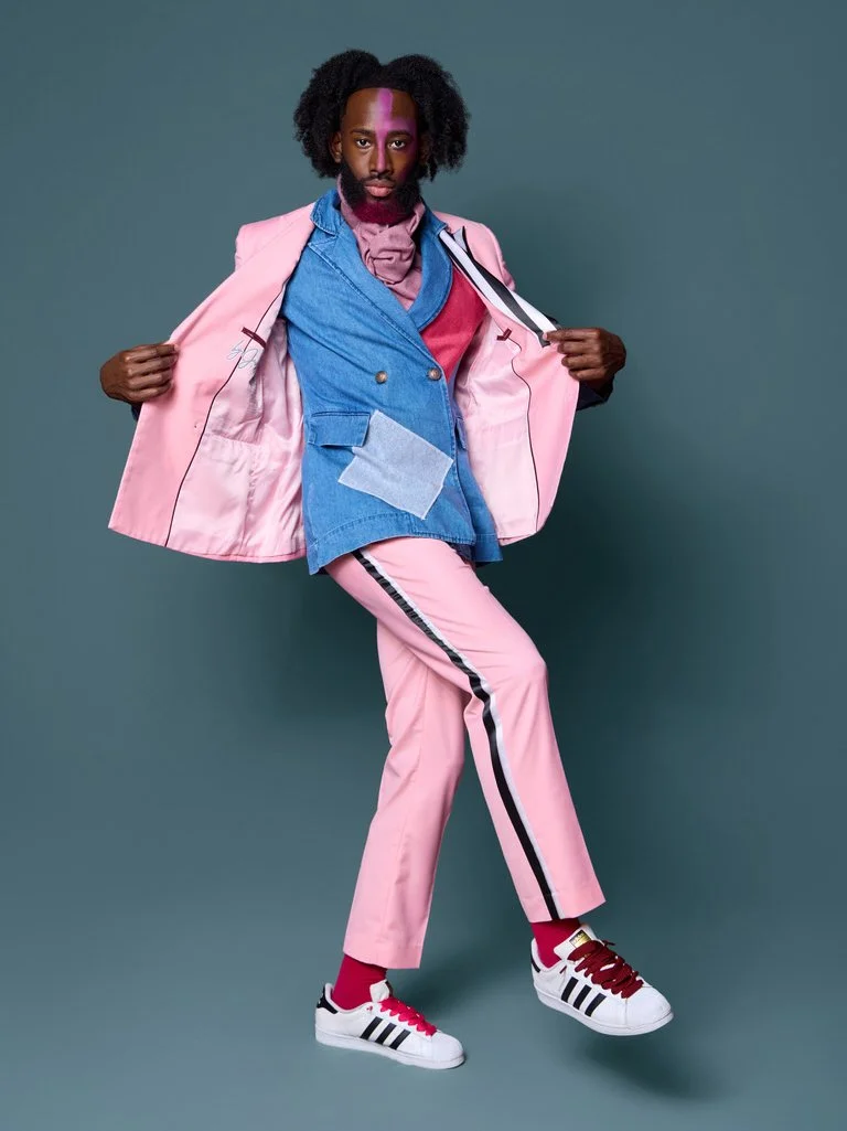 A man with an Afro hairstyle wearing a pink suit, pink pants with black and white stripe, pink shoes with black stripes, a blue blazer, and a pink scarf, posing against a teal background.