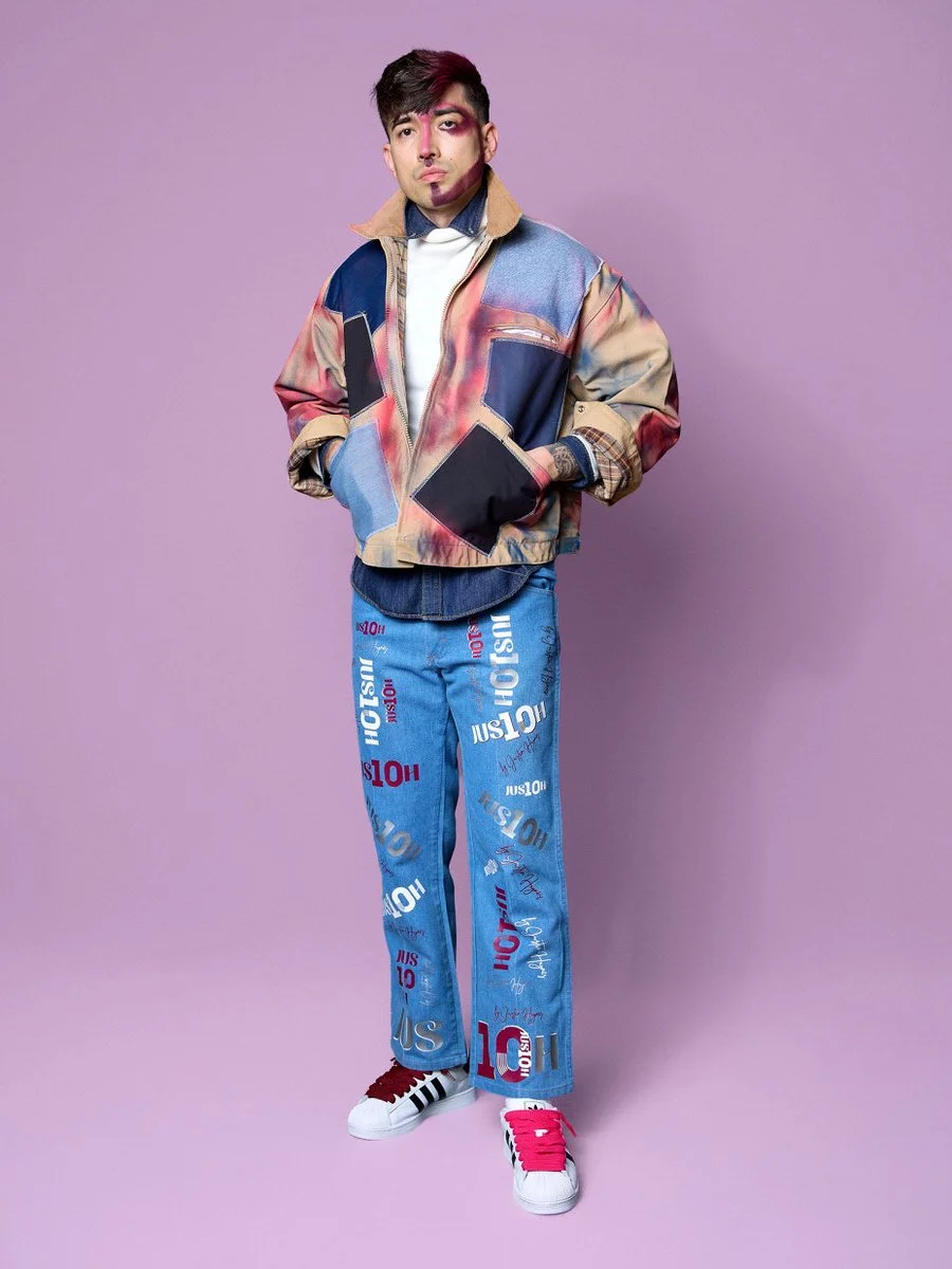 A young man standing against a purple background, wearing a multicolored jacket, branded jeans, and sneakers.