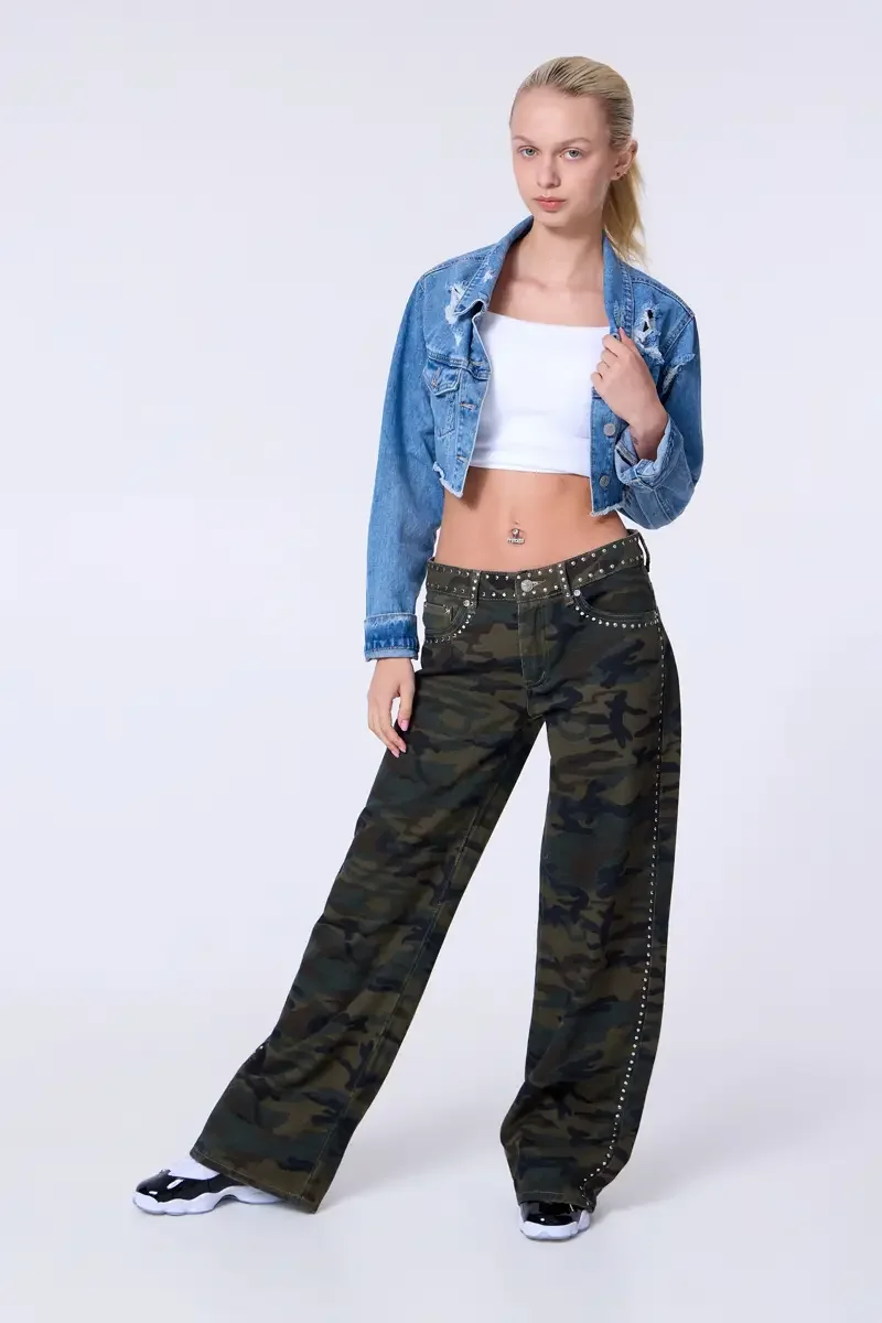 Model portfolio development session, commercial look, cropped distressed blue denim jacket over white crop tank, low-rise studded camouflage wide-leg trousers with silver stud trim at waistband and side seam, full-length, slight turn, one hand raised