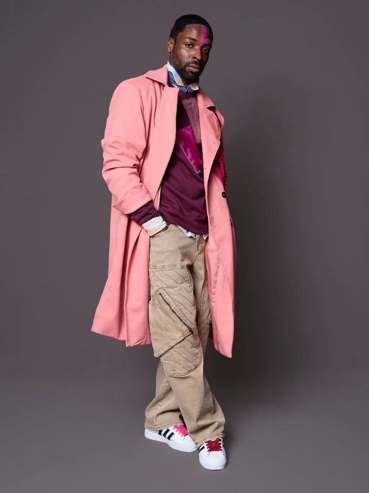 A man standing against a plain gray background, wearing a pink trench coat over layered shirts, beige cargo pants, and white sneakers with red and black accents.