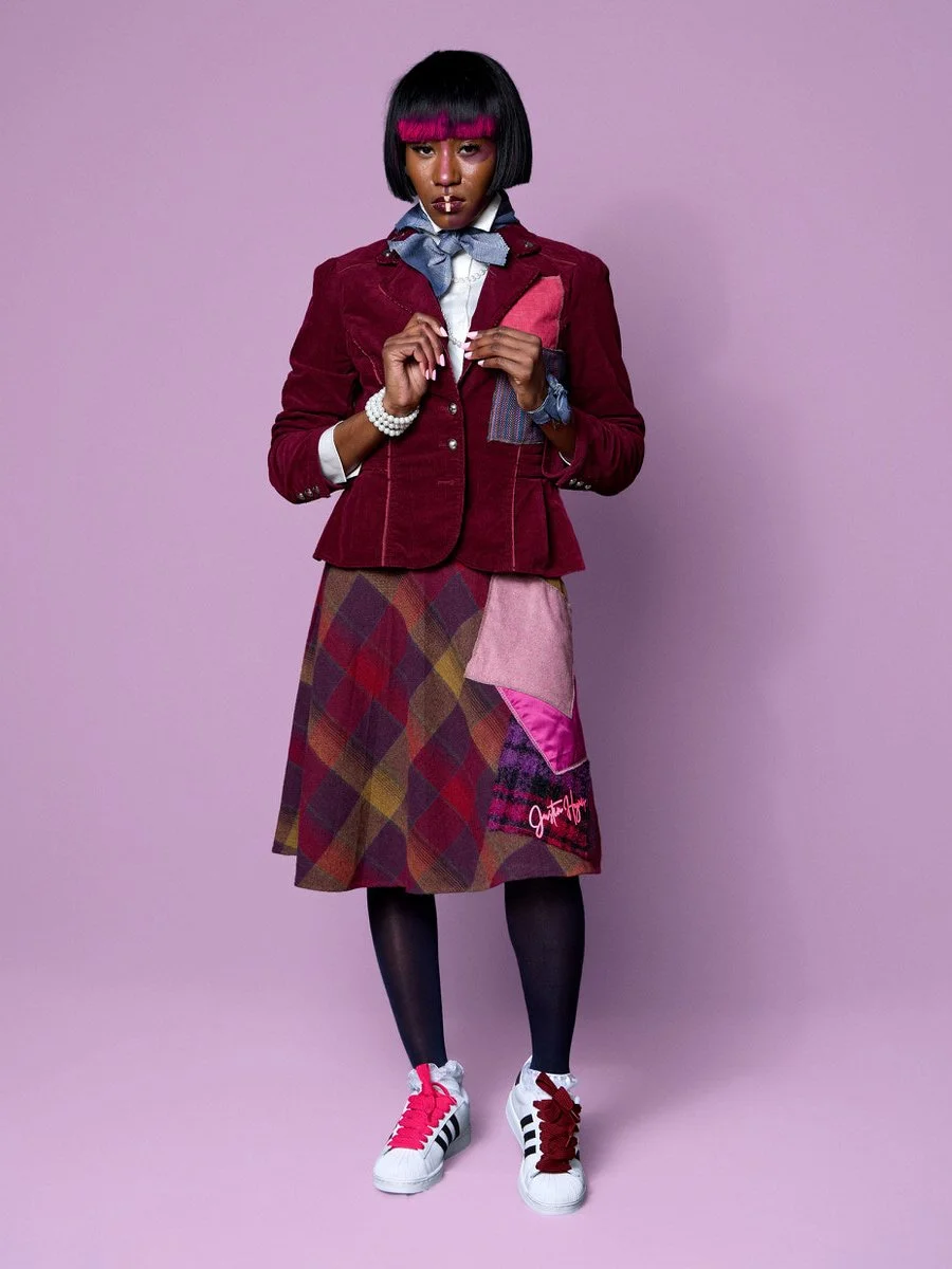 A young woman standing against a pastel purple background, wearing a maroon blazer, layered shirts with a bowtie, a multicolored patchwork skirt, black tights, and white sneakers with red and pink laces. She has a sleek black bob haircut with a pink 