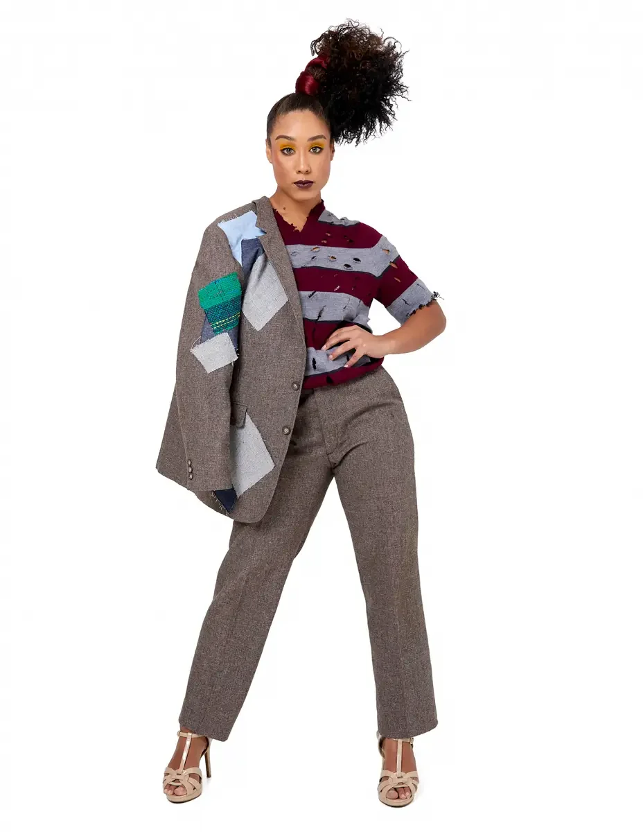 Model portfolio development session, high fashion editorial look, burgundy and heather grey destroyed-knit stripe sweater with raw-edge cutout sleeves, straight-leg heathered trousers, nude t-strap heels, oversized patchwork blazer draped over one ar