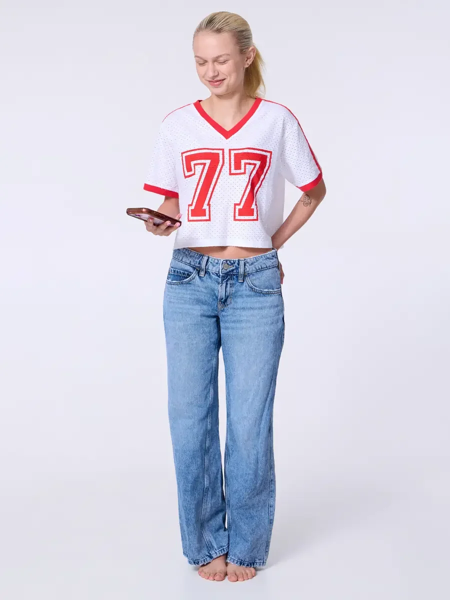 Model portfolio development session, commercial look, white mesh crop football jersey with red number 77 and red trim, low-rise light wash wide-leg jeans, full-length, facing camera, one hand holding phone, gaze downward, soft smile, relaxed stance, 