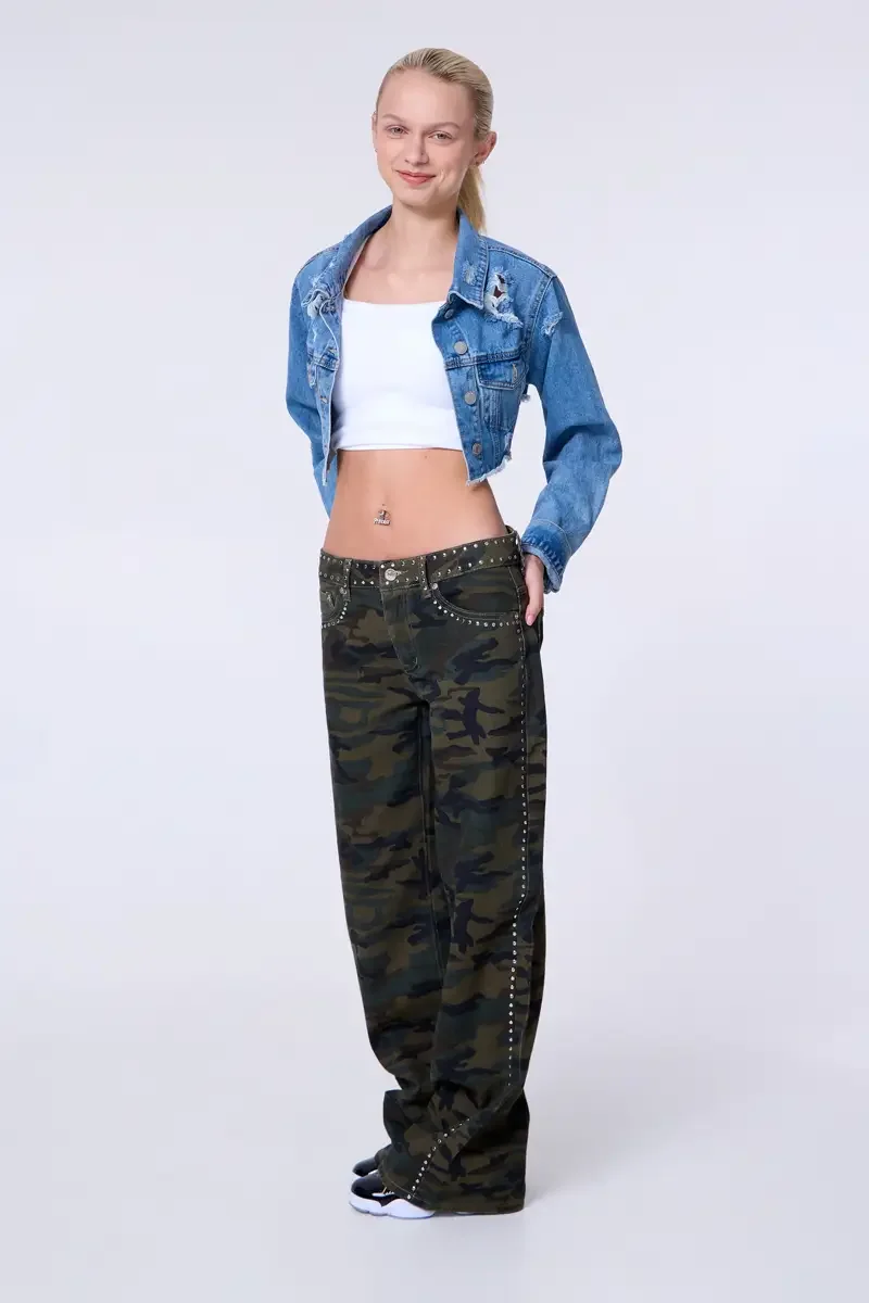 Model portfolio development session, commercial look, cropped distressed blue denim jacket over white crop tank, low-rise studded camouflage wide-leg trousers with silver stud trim at waistband and side seam, full-length, facing camera, one hand in p