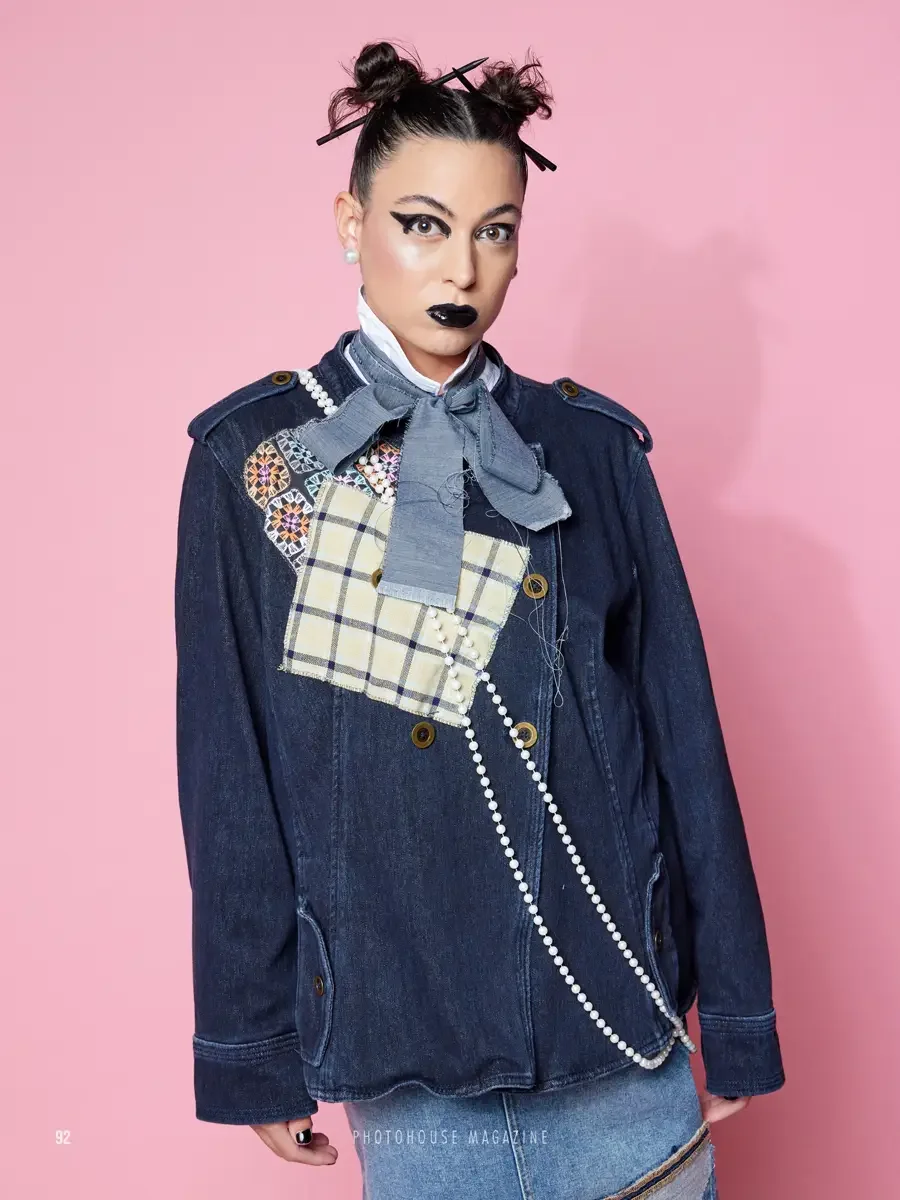 JUS10H 2025 Photohouse Magazine feature, close-up portrait of model in dark navy denim jacket with tartan and multicolor embroidered patchwork panel and grey chambray neck bow, long pearl rope necklace, pink background, photographed by Scott Parker P