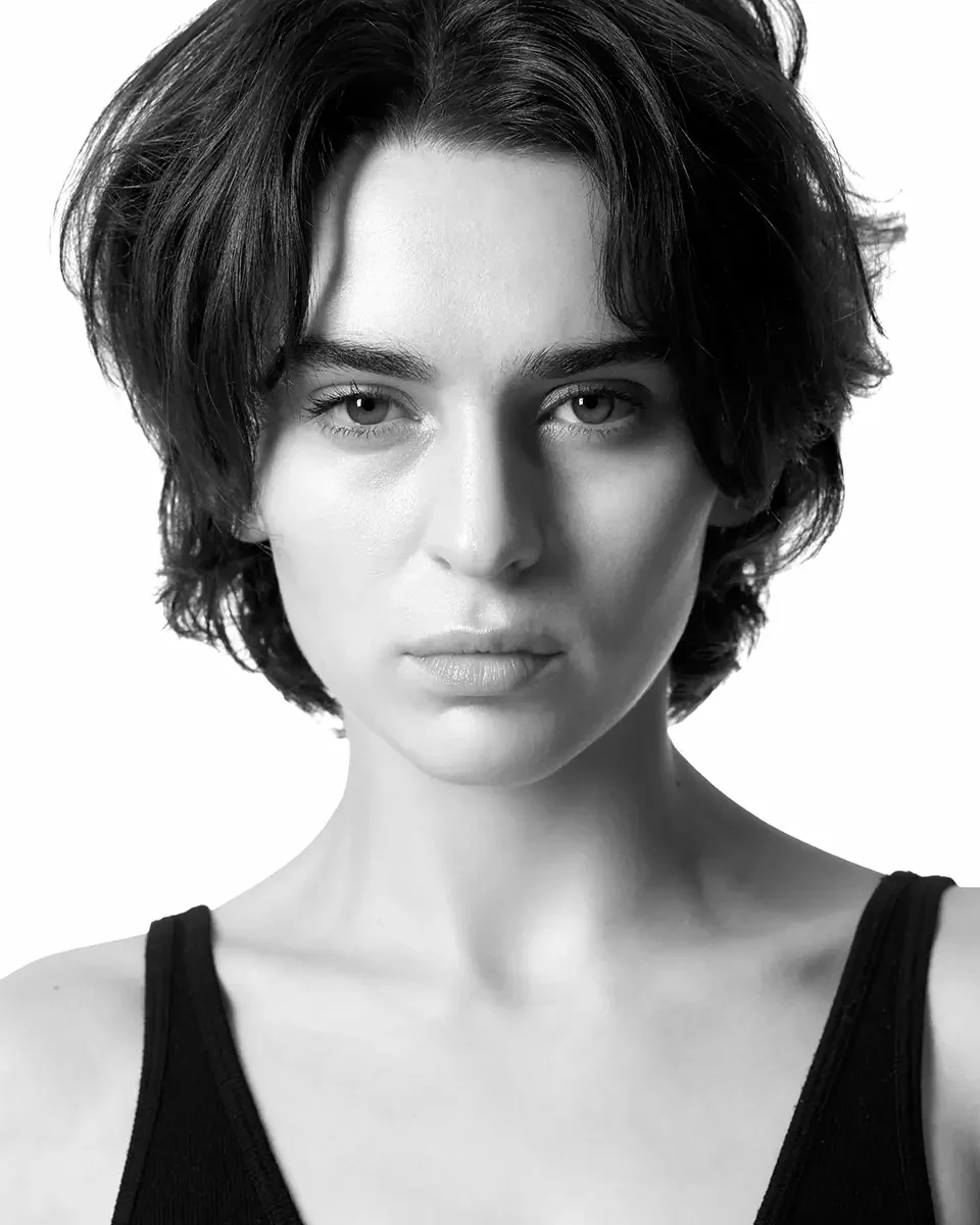 A black and white professional portrait of a model with short wavy dark hair, strong brows, and a natural expression, wearing a black tank top, on a white background, photographed by Scott Parker Photo.