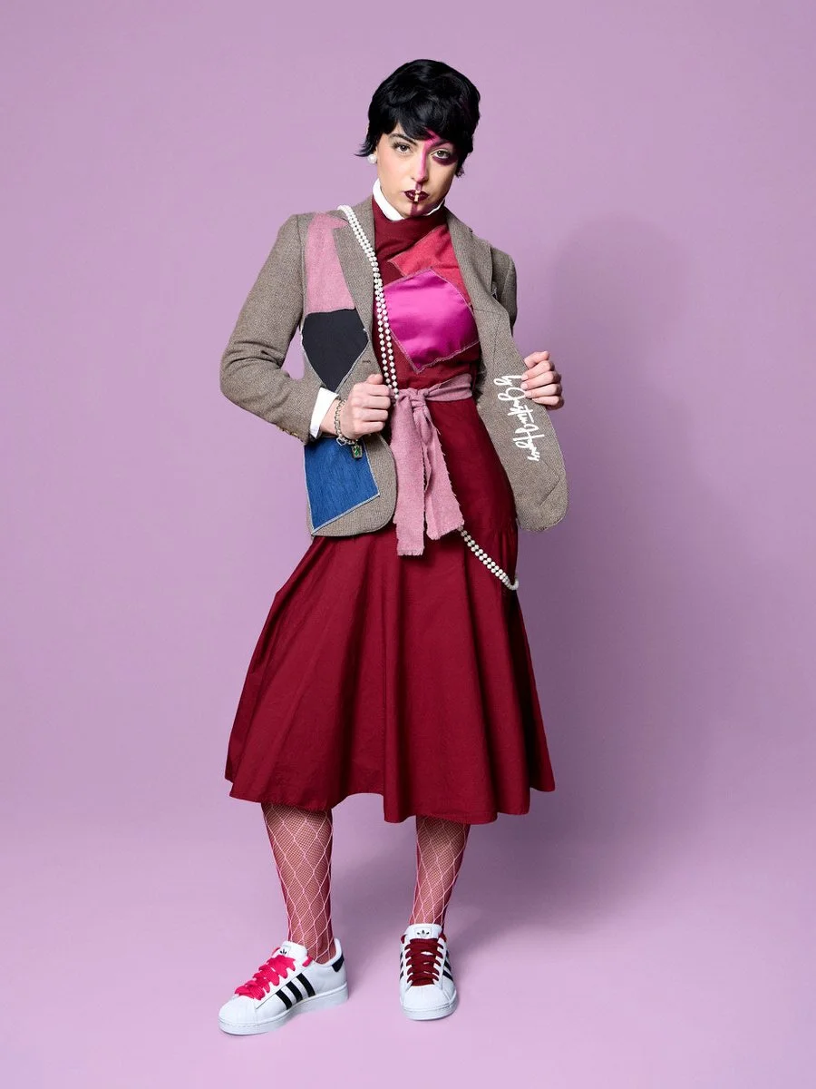 A woman with short black hair dressed in a pink and gray jacket, maroon shirt, and a long red skirt, standing against a pink background. She is wearing white sneakers with red and black accents, fishnet stockings, and holding a pearl necklace.