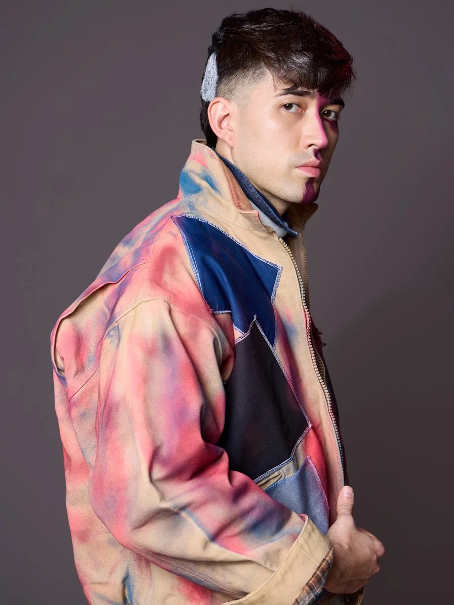 A young man with dark hair and light skin, wearing a multicolored, tie-dye style jacket and posing against a plain dark background.