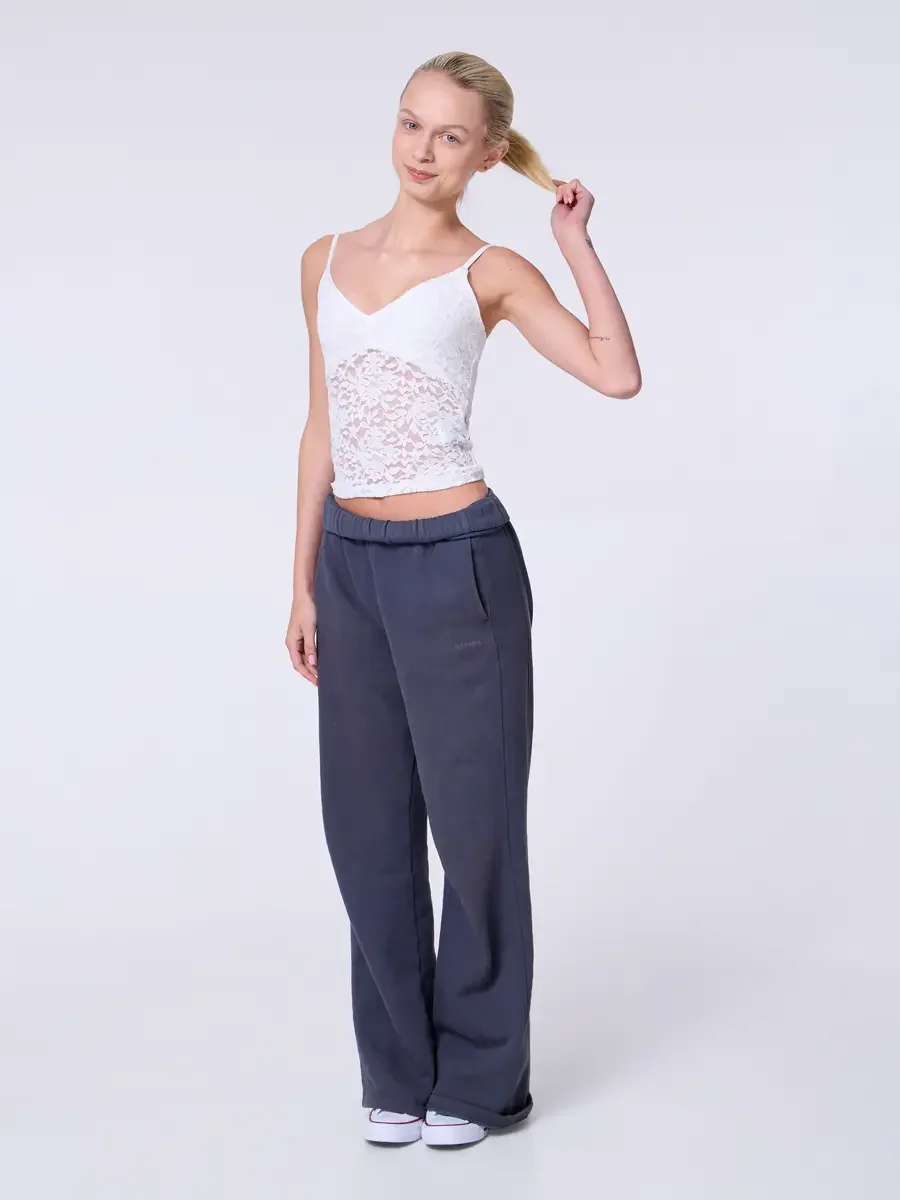 Model portfolio development session, commercial look, white lace crop camisole top and wide-leg charcoal sweatpants with elastic waistband, full-length, facing camera, one hand raised pulling ponytail, relaxed smile, white Converse sneakers, light gr