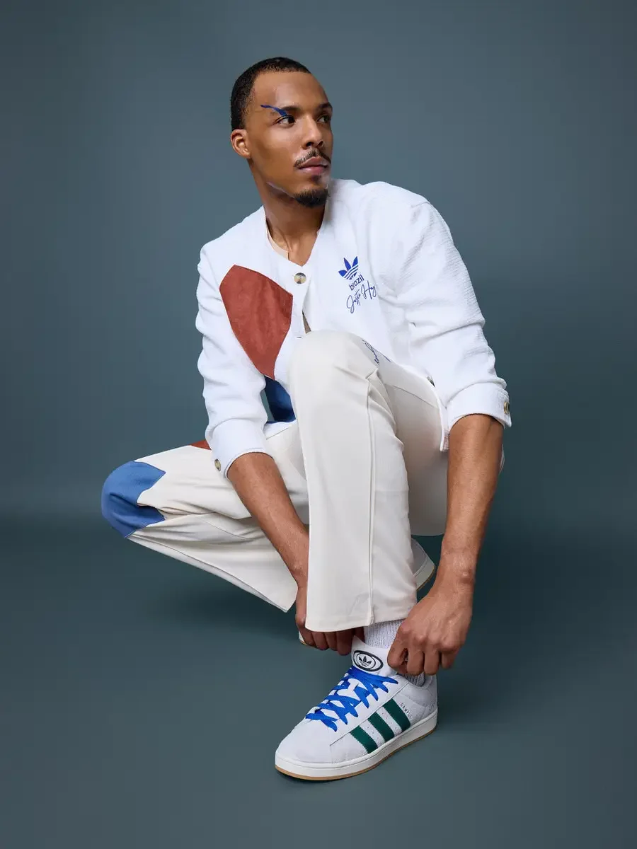 JUS10H x Adidas Brasil campaign, model crouching with gaze upward, white textured patchwork cardigan with terracotta and blue suede panels over cream trousers, Adidas Campus 00s in white with dark green stripes and blue laces, steel blue background, 