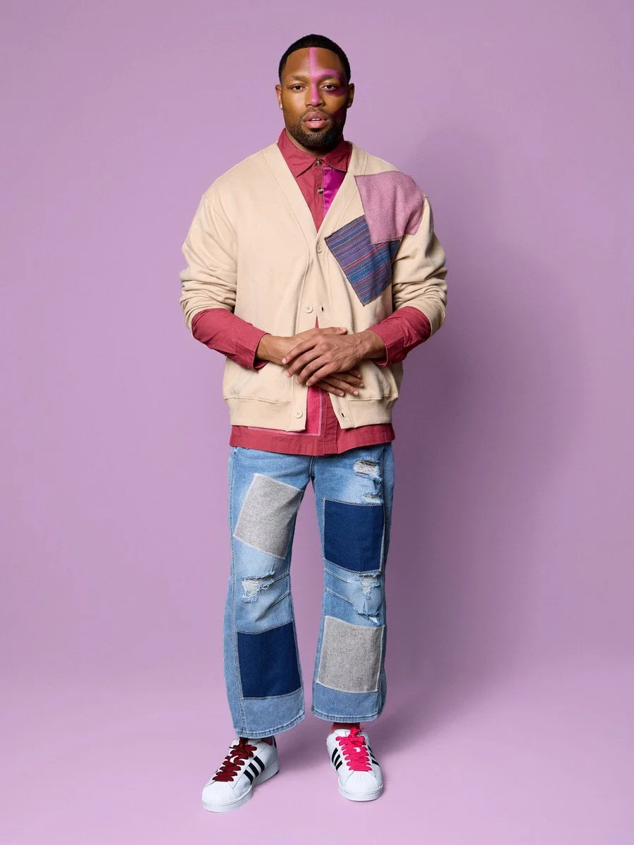 A man standing against a purple background, wearing a beige cardigan with patchwork details over a red shirt, ripped patchwork jeans, and white sneakers with black and red accents.