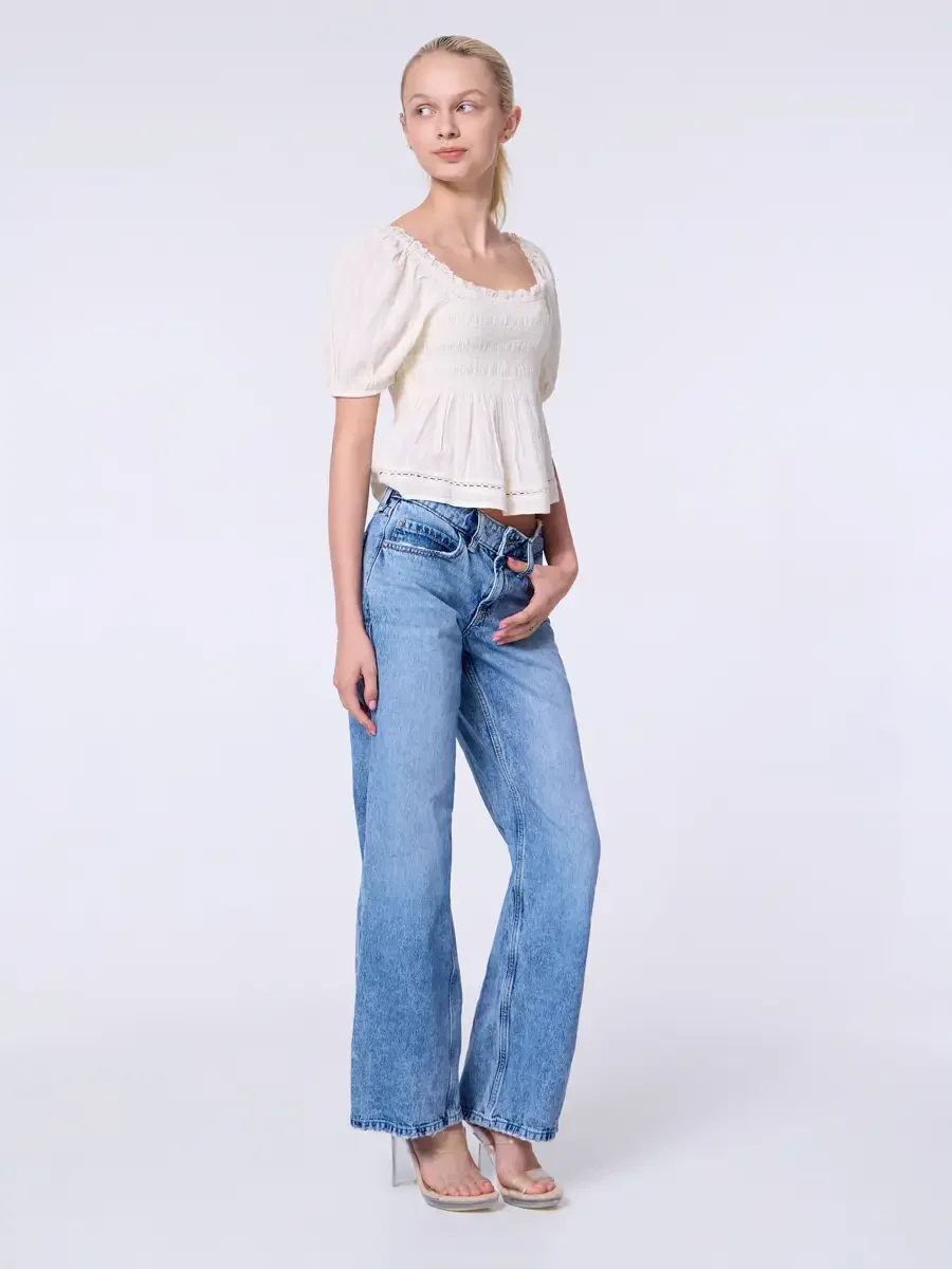 Model portfolio development session, commercial look, cream smocked square-neck puff-sleeve crop top and low-rise light wash wide-leg jeans, full-length, three-quarter turn, one hand in pocket, gaze directed off-camera, elongated posture, nude heeled