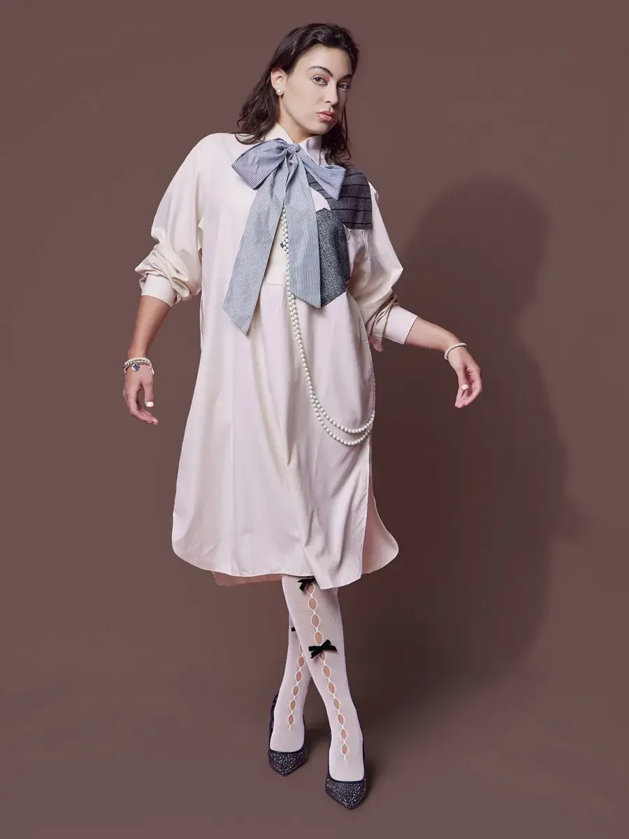 JUS10H 2025 content library, model in motion in blush oversized shirt-dress with striped bow and metallic herringbone patchwork panel, long pearl necklace, bow-detail sheer tights and glitter pumps, brown background, photographed by Scott Parker Phot