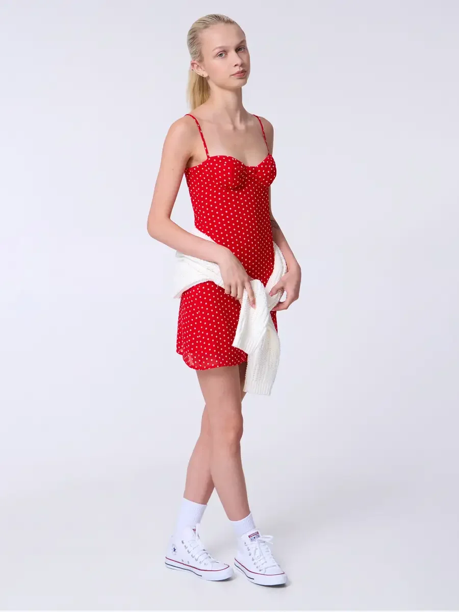 Model portfolio development session, commercial look, red polka dot sweetheart mini dress with cream chunky knit cardigan carried in hand, full-length, three-quarter turn, gaze directed off-camera, cardigan held loosely at side, dress silhouette unob