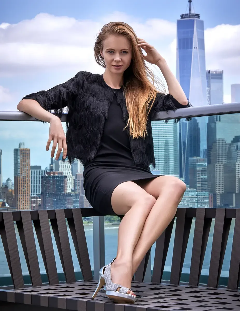 Julia, full length, seated on a rooftop bench with legs crossed, black tiered fringe jacket with zip front, fitted black dress, silver glitter open-toe platform heels, One World Trade Center visible in the Manhattan skyline across the Hudson River, J